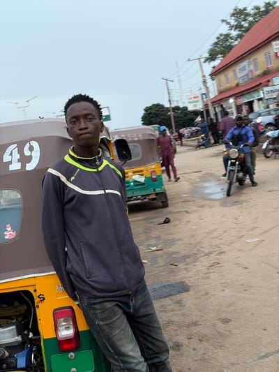 The image captures a street scene in Gura Top, Nigeria, featuring a young man leaning against a brown and yellow auto-rickshaw numbered '49'. He is wearing a dark zip-up jacket with neon green and white accents and blue jeans. Behind him, other auto-rickshaws and motorcycles are visible, along with several individuals walking and riding. Buildings line the street, one with a red roof and signage that appears to indicate businesses. The ground is a dusty, unpaved surface with some puddles and debris. The sky is overcast.