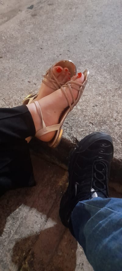 The image captures a close-up, top-down view of two pairs of feet on a textured concrete surface, indicative of an outdoor setting in Juan Griego, Venezuela. On the left, one person wears light gold or champagne-colored strappy sandals with red-painted toenails, paired with black pants. The sandals feature multiple delicate straps across the foot. To the right, another person is seen wearing a black sneaker, identifiable by its distinct rippled design and a visible white Nike swoosh logo on the side, likely a Nike Air Max Plus model. This person is wearing blue jeans. The concrete ground shows variations in tone and texture, with lighter grey areas and darker, possibly shadowed or damp patches. The lighting suggests it is daytime, but with muted sunlight, possibly early morning, late afternoon, or an overcast sky. The image presents a static scene, focusing on the footwear and the ground.