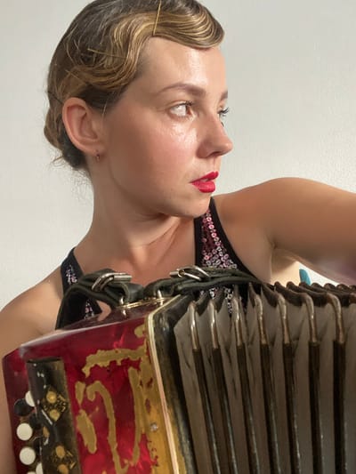 The image shows a close-up, rotated view of a person playing an accordion. The person has fair skin, dark eyelashes, and is wearing bright red lipstick. Their hair is styled with pins, and they have a small earring in their left ear. They are wearing a black, sequined top with straps. The accordion is red with gold detailing and black bellows. The scene appears to be indoors, against a plain white background. The lighting suggests it could be daytime. There is no visible text in the image. The context of Podgorica, Montenegro, is provided but not directly discernible from the image itself.