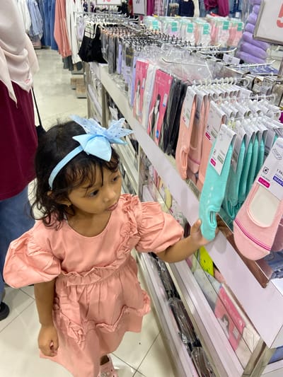In Sidoarjo, Indonesia, a young girl with curly black hair and a blue polka dot bow headband is browsing socks in a retail store. She is wearing a pink puffy-sleeved dress with ruffles. She reaches out to touch a pair of mint green no-show socks hanging on a display rack. The store is well-lit with rows of clothing and accessories displayed on shelves and hanging racks. Other shoppers are visible in the background, and clothing items are hung on racks in the distance.
