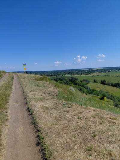 The image depicts a rural landscape under a clear blue sky with scattered white clouds. In the foreground, a dirt path winds its way up a grassy hill, with dry, straw-colored grass dominating the slope. Further up the hill, a flag pole stands with two flags: a blue and yellow flag (likely the Ukrainian flag) and a yellow flag below it. Beyond the hill, a sprawling green valley unfolds, dotted with trees and a village of houses. A few winding paths are visible in the valley floor. The scene suggests a serene, natural environment.