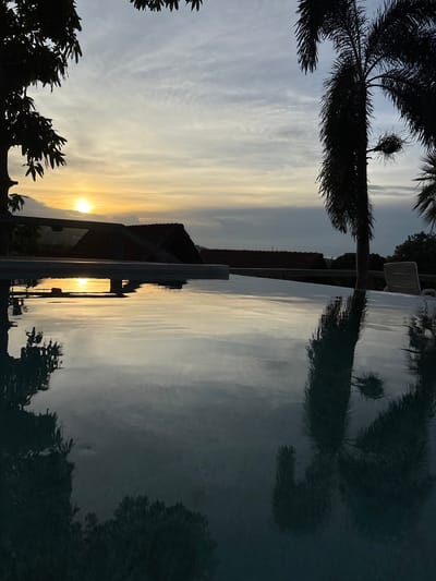 The image depicts a serene sunset scene at an infinity pool, likely located in Ban Bang Rak, Thailand. The sun, a brilliant orb of light, is low on the horizon, casting a warm glow across the sky and reflecting on the pool's surface. Silhouetted against the vibrant sky are the dark shapes of palm trees and foliage, framing the view. In the background, terracotta-tiled roofs of buildings are visible, suggesting a residential or resort setting. The water of the infinity pool creates a mirror-like surface, reflecting the sky, trees, and surrounding environment with remarkable clarity, enhancing the overall tranquility of the scene. There are no people or animals visible in the image. The setting is peaceful and picturesque, capturing the essence of a tropical evening.