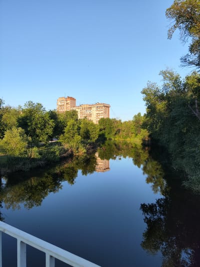 The image captures a serene scene of a calm body of water reflecting a clear blue sky and lush greenery. In the background, two large, multi-story buildings stand tall amidst a dense arrangement of trees. The buildings appear to be residential, with numerous windows visible on their facades. The trees form a thick, verdant line along the water's edge and extend into the distance, creating a natural barrier between the urban structures and the water. The water itself is still, creating a near-perfect mirror image of the surrounding landscape and the sky above. In the foreground, a white railing runs along the bottom of the frame, suggesting the viewpoint is from a bridge or elevated walkway overlooking the scene. The overall atmosphere is tranquil and picturesque, with the soft lighting indicating either early morning or late afternoon. This scene is typical of the natural areas found within the city of Kryvyi Rih, Ukraine, where urban development often coexists with accessible green spaces and waterways like the Inhulets River or its tributaries.