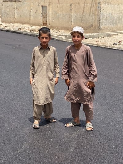Two young boys are standing on a paved road in what appears to be a city or town setting. The boy on the left wears a beige-colored shalwar kameez and a black skullcap. The boy on the right is dressed in a brown shalwar kameez and a light-colored hat. Both are wearing sandals. The background features a wall or building, and the scene is bathed in bright daylight, suggesting it is outdoors. The exact location is not clear, but the prompt suggests Kandahar, Afghanistan.