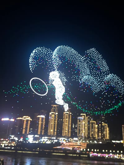 This image captures a nighttime drone show over the cityscape of Jiangbeicheng Street, China. Numerous white and colored lights form abstract shapes in the sky, with a prominent white humanoid figure and a ring shape being particularly noticeable. The city skyline, illuminated by the lights of numerous tall buildings, stretches across the lower portion of the frame, with a body of water reflecting the urban glow in the foreground. The sky is dark, serving as a canvas for the illuminated display.
