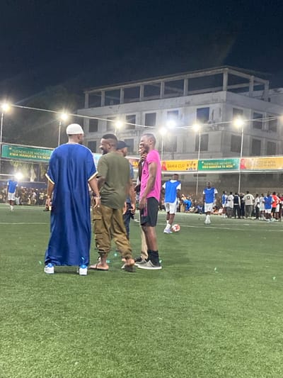 The image shows a group of five men standing on a green artificial turf soccer field at night, located at the coordinates -6.764, 39.258 in Dar es Salaam, Tanzania. The men are interacting and smiling. The man in the blue robe is wearing a white skullcap and is positioned to the left of the frame, while the other four men stand to his right. The man in the pink t-shirt is standing to the far right of the group. The others are wearing casual attire. The man in the green shirt is wearing cargo pants. They are standing in front of a fence with lights overhead. There is a sign in Swahili on the fence and the word "SAMA CUP" is also visible. Several more people are visible in the background, blurred, watching a soccer game on the field. There is a soccer ball on the field to the right. The scene is lit by overhead lighting. The image is taken at night.
