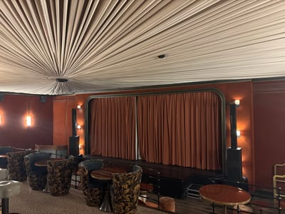 The image depicts an empty, intimate screening room or small theater, identified as the Club de l'Étoile located at 14 Rue Troyon, 75017 Paris, France. The scene features a raised stage positioned at the front, equipped with closed, rust-colored velvet curtains. The stage platform is dark, likely black or a dark wood finish. Flanking the stage are tall, black, columnar structures that incorporate vertical lighting elements, illuminating the adjacent, reddish-brown upholstered walls. Additional wall sconces with two exposed bulbs each provide ambient, warm lighting throughout the room.
The ceiling presents a distinctive design, with light-colored, pleated fabric panels radiating outwards from a central circular fixture, creating a sunburst or fan-like pattern. The seating arrangement consists of multiple dark, ornate armchairs with floral patterns, positioned around small, round, dark wood tables with metallic bases. On the far left, a few lighter-colored, upholstered stools or low chairs with metallic bases are also visible. The floor is covered with a light-colored, speckled carpet. No individuals are present, and no actions or interactions are occurring.