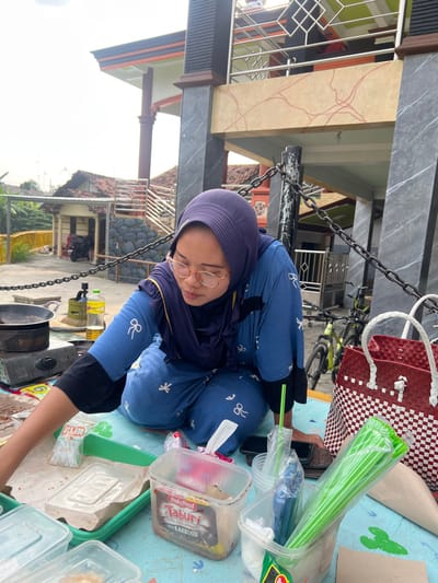 A female individual, wearing a dark blue hijab, light blue long-sleeved shirt with white bow motifs, and matching pants, is seated on a light blue mat decorated with white cactus patterns. She wears light-rimmed glasses and is observed looking down, her right arm extended towards various plastic food containers and utensils. One clear plastic container prominently displays an "Indofood TABURI SAUS BARBEKIU" label. To her left, a portable gas stove with a black pan is positioned on the mat, alongside a yellow bottle of cooking oil and a small gas canister. A red and white woven plastic bag, a black mobile phone, and a brown wallet are also present on the mat near her. In the background, a two-story building with dark grey marbled pillars and beige panels featuring red linear patterns is visible, complete with a balcony and metal railing. A black metal chain fence spans the midground. Beyond the fence, several residential buildings with terracotta-tiled roofs, green bicycles, and distant vegetation are discernible under an overcast sky. This scene is situated in Kecamatan Pati, Indonesia.