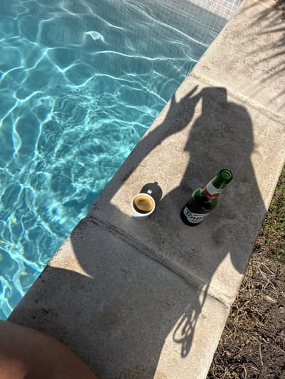 The image depicts a poolside scene in Antibes, France. In the foreground, a concrete poolside edge borders a turquoise swimming pool. On the concrete, a small white cup filled with dark liquid, likely coffee, sits next to a green glass bottle of "Tourtel" beer. The shadow of a person's hand making a peace sign is cast on the concrete, along with the shadow of the beer bottle and the person's head and torso. The water in the pool is clear and exhibits light reflections from the sun. The background shows a small patch of dry grass and soil. The overall impression is of a relaxed, sunny day by the pool.