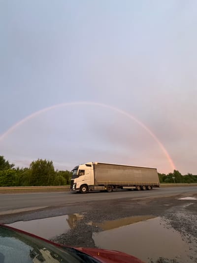 This media file depicts a truck driving on a road in Sovkhoznyi, Russia. A faint rainbow is visible in the sky, suggesting recent or ongoing rain. The road appears wet in places, with puddles reflecting the overcast sky and surrounding environment. Trees line the side of the road, and a red car is partially visible in the foreground, suggesting the photo was taken from another vehicle. The overall lighting indicates it is likely late afternoon or early evening. The image focuses on the truck and the natural phenomenon of the rainbow, creating a serene and somewhat moody atmosphere. There is no visible text in the image.