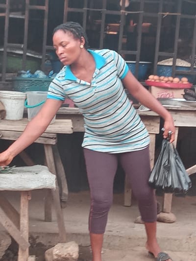 A medium shot captures a Black woman wearing a striped polo shirt and leggings standing at a market stall in Sabon Tasha, Nigeria. The woman has braided hair and is looking downwards and to her left. Her right hand is extended, holding a black plastic bag. The market stall features wooden structures and tables laden with various goods, including buckets and a red tray filled with small, round items, possibly food. The ground appears to be dirt. The background shows a metal grate structure, common in market environments.
