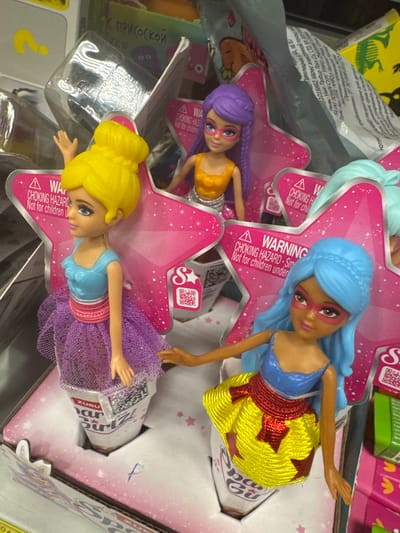 This media file shows three dolls in their packaging, likely in a retail setting. The dolls have distinct features: one has purple hair and an orange top, another has blue hair and a yellow skirt with red stars, and the third has blonde hair and a purple sparkly skirt. All dolls have a warning label indicating "CHOKING HAZARD - Not for children under" in English. There are also Russian words visible, such as "ПРИСОСКОЙ" which translates to "with a suction cup", suggesting some of the toys may have this feature. The packaging features pink stars and glitter. The overall scene appears to be a shelf display in a store, possibly a toy store. The lighting is artificial, consistent with an indoor setting. The presence of Russian text suggests the location could be in Russia. The dolls appear to be part of a toy line, with "ZURU" and "Star Girl" visible on some packaging, indicating the brand and product name. There are no clear indicators of time of day or weather.