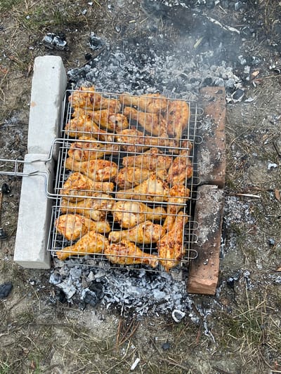 In Stara Rafalivka, Ukraine, a barbecue grill containing several rows of cooked chicken pieces rests on a bed of grey ash and embers. The grill is supported by two concrete blocks on the left and a brick on the right. The chicken pieces, appearing to be drumsticks and wings, are golden brown and appear well-cooked. Wisps of smoke rise from the grill, indicating it is still actively being used or has recently been used. The surrounding ground is covered with dry grass, twigs, and scattered pieces of charcoal, suggesting an outdoor setting. The lighting suggests it is daytime, possibly overcast.