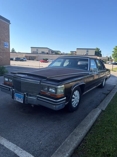 The image shows a dark-colored Cadillac parked on an asphalt surface next to a concrete curb. The car has Illinois license plate number CU 22756. In the background, buildings are visible, and a clear blue sky is overhead. The location appears to be in a parking area. The address is not provided, but the license plate indicates the location is in Illinois, which would include the city of Morton Grove, United States of America.