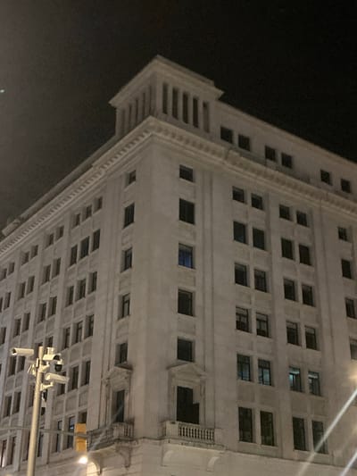 The image shows the facade of a large, light-colored building at night in Barcelona, Spain. The building has a classical architectural style with rows of windows and decorative elements. A street light is visible on the right side of the image, and a pole with multiple security cameras is on the left. The sky is dark and there is a faint light in the upper left quadrant.