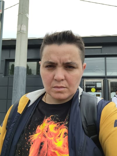 A person with short, dark hair is standing outdoors in front of a modern building with dark gray siding and large windows. They are wearing a black t-shirt with a fiery horse graphic, a navy and yellow jacket, and a gray backpack. A lamppost with stickers is to the left of the person. The background includes reflections in the windows of trees and what appears to be a street scene. The overall lighting suggests an overcast day. The location is identified as Ramenskoye, Russia.