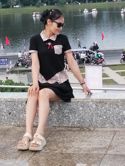 In Da Lat, Vietnam, a young woman with dark hair pulled back in a bun and wearing sunglasses is seated on a granite ledge, looking to her right and downward. She is dressed in a black top with a plaid collar and sleeve trim, a decorative red bow on the neckline, and a black skirt. Her feet are adorned with light-colored wedge sandals. In the background, a body of water with swan-shaped boats is visible, with a pathway and road beyond, where people are walking and riding motorbikes. A red flag stands near the road. The overall setting appears to be a park or public area.
