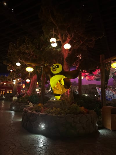 An outdoor nighttime scene depicts a large, illuminated statue of Po, the panda from Kung Fu Panda, striking a martial arts pose. Po is yellow and green, with a prominent belly. He stands on a circular stone pedestal surrounded by lush greenery. Behind him, a large, gnarled tree is adorned with numerous paper lanterns of various sizes and colors, including white, yellow, and red. Some lanterns are lit, casting a warm glow. The background features a stylized, mountainous landscape with purple and pink hues, illuminated by more lanterns and possibly artificial lighting. To the left of the tree, traditional Chinese architecture, including red buildings with ornate windows, is visible. A person is seated on the stone pedestal to the left of Po, looking at their phone. The ground is paved with large, irregular stones. The overall atmosphere is festive and illuminated, suggesting a themed area or attraction within a city like Tongzhou District, China, known for its entertainment venues.