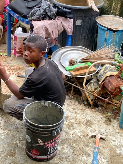 Ado, Nigeria, shows a person in a black shirt and light trousers kneeling next to a black bucket with a silver rim that has the word "Finec" on it. The ground is cement-like. In the background, there are items stacked in an untidy fashion including blue plastic containers, a metal bucket, a broom, a level, and a rusty metal object. A hammer with a blue handle is lying on the ground nearby. Clothes, a black bag, a box of facial tissues and other personal items are situated near the edge of the frame.