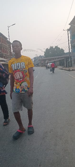 The image displays a street scene in Aba, Nigeria, under diffused daylight. In the foreground, a young male with dark skin and short dark hair stands slightly left of center, facing the camera. He wears a yellow short-sleeved t-shirt with a "DIOR" logo, a cartoon fist, and other colorful characters, along with light grey shorts and dark slip-on clogs featuring pink straps. To his left, the lower body of another person, wearing dark trousers and a dark sandal, is partially visible. The paved road recedes into the distance, occupied by pedestrians and vehicles. A person in a red shirt walks away from the camera in the midground, carrying a dark object. Multi-story buildings with balconies line the left side of the street, while utility poles with overhead wires and a street lamp are visible against a light, hazy sky.