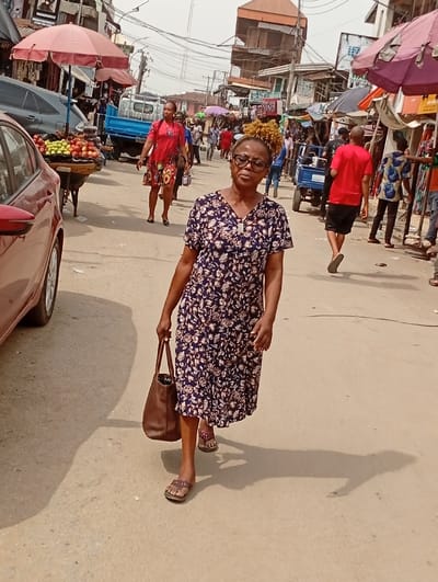 A woman wearing a floral dress and sandals walks toward the camera, holding a brown leather bag. The scene is a busy street, likely a market, with various vendors and pedestrians. There's a red car on the left, and fruit is displayed on a cart nearby. Other people are visible further down the street, some under umbrellas. Buildings and overhead wires fill the background, creating a bustling urban environment. The light suggests a sunny day.