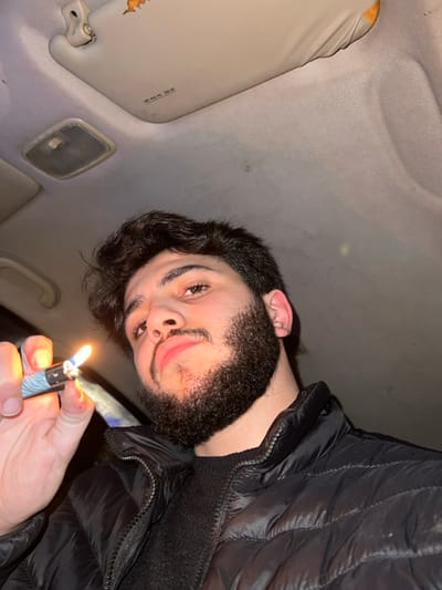 A selfie taken from a low angle inside a car shows a bearded man holding a lit lighter. The car's interior is visible, including the headliner, a visor, and an overhead light. The man is wearing a black, quilted jacket. The light from the lighter illuminates his face and hand. The overall lighting is dim.