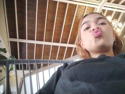 The image is a selfie taken indoors, possibly in a restaurant or cafe. A young woman with reddish-brown hair, wearing a dark gray sweatshirt and large hoop earrings, is puckering her lips for the camera. She has full lips with pink lipstick. The background shows a wooden ceiling with white structural beams and a decorative chandelier. A railing, possibly of a balcony or staircase, with gray cords threaded through it, runs across the lower portion of the image. A plant is partially visible on the left. The location is identified as Porlamar, Venezuela.