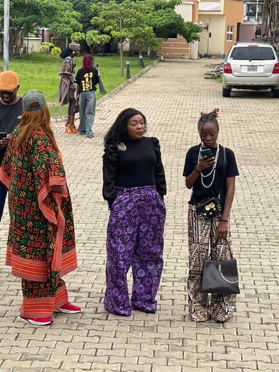 In Barakin Lerawi, Nigeria, three women are standing on a paved outdoor area. The woman on the left is wearing a vibrant, multi-colored patterned kaftan with orange trim and red sneakers. She is looking down at her phone. In the center, a woman is wearing a black top and high-waisted purple pants with a floral pattern. She is looking to her right. To the right, a young woman is wearing a black t-shirt, a long pearl necklace, and patterned pants. She is looking down at her phone and holding a black handbag with a gold chain. In the background, a silver SUV is parked on the right side of the frame. Trees and foliage are visible behind the women and the vehicle. The area appears to be a driveway or courtyard with a cobblestone-like pavement.