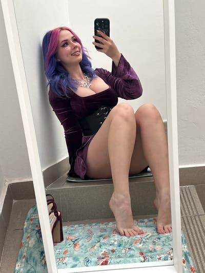 A young woman takes a photo of herself in a mirror. The woman has long hair dyed purple and blue, wears a dark purple long-sleeved robe-like garment over a black corset and a pearl necklace. Small heart stickers adorn her cheeks. She holds a black smartphone in her left hand. The mirror reflects a view of her legs and feet, which rest on a surface covered with a patterned turquoise rug. Next to her feet are a pair of burgundy high heels. The scene appears to be in a bathroom with tiled walls. This image does not appear to be located in the city of Zagreb, Croatia.