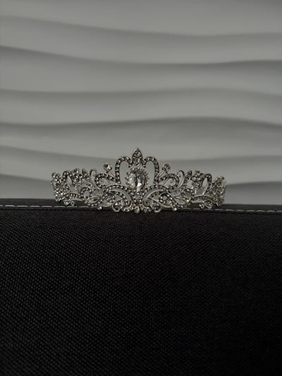 The image shows a close-up, side-view of a silver tiara adorned with rhinestones. The tiara is placed on the edge of a dark, textured surface, possibly a piece of furniture or fabric, which takes up the right half of the frame. The background on the left is a white, undulating surface, creating a subtle ribbed pattern with shadows and highlights, suggesting a decorative wall or paneling. The lighting is soft, casting gentle shadows and highlighting the intricate details of the tiara. The focus is sharp on the tiara, with the background elements softly blurred, drawing attention to the decorative piece. There is no visible text, people, or indication of a specific time of day or weather. The overall impression is one of elegance and delicate craftsmanship.