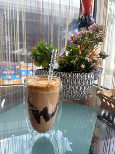 A glass of iced coffee sits on a glass table. The coffee is layered with milk and appears to have chocolate syrup swirled in it. A clear straw protrudes from the drink. In the background, a potted plant with small pink flowers and green leaves is visible. Next to the plant is a decorative vase with a textured, patterned surface. The scene appears to be indoors, near a window with blinds that cast horizontal shadows. Outside the window, a cityscape is faintly visible, suggesting an urban setting. The lighting suggests it could be daytime, possibly late morning or afternoon. No specific emotions are conveyed, and no text is legible in the image. The overall impression is a casual moment, enjoying a refreshing drink in a bright, indoor environment.