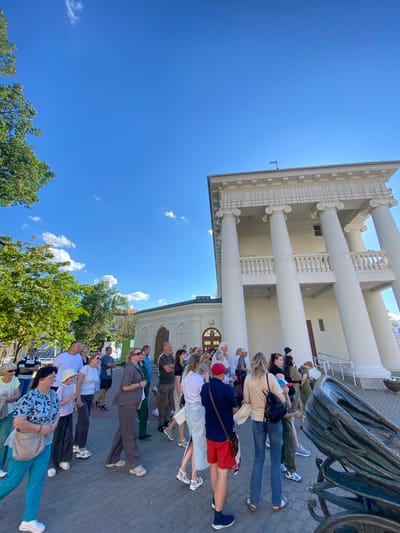 A group of people are gathered outdoors in front of a building with large white columns. They are all facing the same direction, appearing to be listening to someone or observing something. The building has a classical architectural style. The sky is clear and blue, suggesting a sunny day, likely late morning or early afternoon given the brightness. Some of the people are dressed casually, while others appear to be in more formal attire. There is a large, ornate object, possibly a historical artifact or sculpture, in the foreground. The ground is paved with cobblestones. The atmosphere appears relaxed and attentive. There is no visible text in the image.