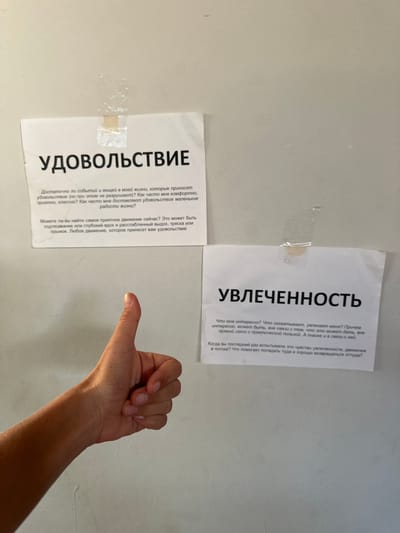 A human arm and hand is visible in the foreground of this image, with the hand in a thumbs-up gesture. The arm and hand are positioned in the lower right quadrant of the frame, casting a shadow on the light-colored wall behind it. Two sheets of paper are affixed to the wall with tape. The paper on the left is titled "УДОВОЛЬСТВИЕ" (Pleasure) and contains text in Russian asking questions about enjoyment and life's small joys. The paper on the right is titled "УВЛЕЧЕННОСТЬ" (Engagement/Enthusiasm) and also contains Russian text, posing questions about interests and the feeling of being in a flow state. The lighting suggests an indoor setting, possibly during the day. The overall impression is one of contemplation or self-reflection, prompted by the questions on the posters.