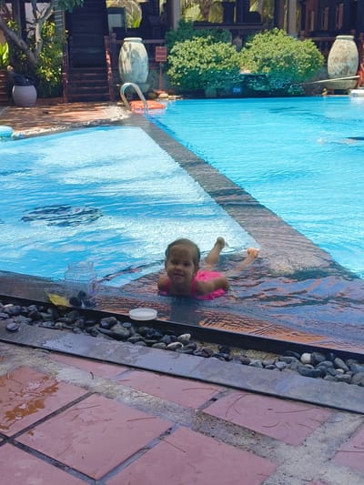 This media file captures a candid scene at a swimming pool. The primary subject is a young child, likely a toddler, wearing a pink swimsuit. The child is positioned near the edge of the pool, with their body submerged in the shallow water and their head and upper chest visible above the surface. They are looking directly at the camera with a faint smile.

The setting is an outdoor swimming pool with clear blue water. The pool has a dark, textured edge, and beyond that, there are smooth, reddish-brown tiles on the deck, some of which are wet and reflective. A border of dark, smooth stones lines the edge of the pool. In the background, there are lush green bushes and trees, suggesting a tropical or subtropical location. Further back, architectural elements like wooden structures and large decorative urns are visible, hinting at a resort or hotel setting.

A clear glass jar with dark objects inside and a yellow leaf is placed on the pool edge near the child. A white lid is also visible on the pool edge. The water is calm, with ripples reflecting the sky.

The lighting suggests it is daytime, possibly late morning or afternoon, with the sun casting reflections on the water. There are no clear indicators of specific weather conditions beyond the presence of water and sunlight. The overall mood appears relaxed and joyful, with the child's expression conveying a sense of contentment.

A sign with the text "2m" is partially visible on a green bush in the background, indicating the depth of the pool in that area.

While the precise location of Phu Thuy Ward, Vietnam is provided as context, there are no overt visual cues in the image that specifically identify it as such, other than the general impression of a Southeast Asian tropical environment.