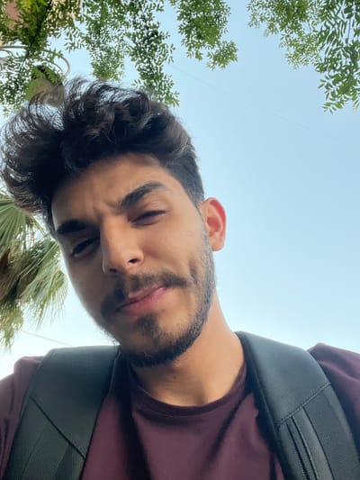 An outdoor, low-angle portrait captures a young man with dark, wavy hair and a short beard, looking directly down towards the camera. He is wearing a dark maroon t-shirt, and a black, textured backpack strap is visible on his right shoulder. His eyes are partially closed or looking downwards, and his lips are slightly parted in a relaxed expression.
The background is dominated by a clear, bright blue sky, suggesting daytime with good visibility. Green foliage from trees frames the upper left side of the image, while a palm frond is visible on the lower right. A thin, dark line, possibly a wire, stretches diagonally across the upper portion of the sky. The overall setting appears to be a sunny outdoor environment, consistent with Al-Muhajerin Municipality, Syria. There is no visible text in the image. The man's demeanor appears neutral and casual.