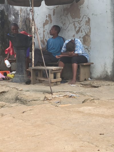 The image shows two individuals, one man and one woman, seated outside a building with a distressed, cracked white wall in Aba, Nigeria. The man is wearing a blue shirt and black pants, while the woman is wearing a blue and white patterned shirt and black pants. Both are seated on small wooden stools. An old water pump with a handle is located nearby. Various objects such as a plastic container and other miscellaneous items are scattered around the base of the pump. The ground appears to be concrete, cracked, and showing signs of wear.