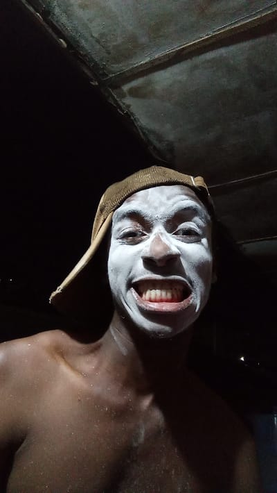 A dimly lit portrait features a young individual with dark skin, captured in a close-up, head-and-shoulders shot. The person is looking directly at the camera with wide, expressive eyes and a broad, toothy grin, revealing both upper and lower teeth. Their face is covered in a thick, white substance, possibly paint or a mask, which starkly contrasts with their natural skin tone and highlights their features. A brown, textured cap is worn on their head. The individual appears shirtless, with their shoulders and upper chest visible, showing a light sheen on the skin, possibly from moisture or the applied substance.
The background is predominantly dark, suggesting either a nighttime setting or a poorly lit indoor space. Above the person's head, a dark, textured surface, possibly a ceiling or roof, is partially visible, indicating a sheltered environment. The artificial lighting, likely a flash, illuminates the subject's face prominently. The person's exaggerated smile conveys a sense of cheerfulness, excitement, or playfulness. This image, possibly taken in Onitsha, Nigeria, could depict participation in a local event, a costume, or a moment of whimsical self-expression. No text is visible in the media.