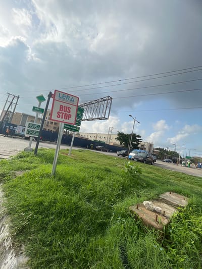 The image shows a bus stop sign in Lekki Peninsula Phase 1, Moba, Nigeria. The sign has the logo of LERA (Lekki Peninsula Residents Association) and states "BUS STOP" in large letters. Below the bus stop sign are signs indicating prices for distributors and retailers, displaying "0000:0" and "250:0", respectively. There are also other signs with text that is partially obstructed. In the background, there is a street with cars parked along the roadside. Lush green grass borders the street. There are utility poles with power lines visible in the sky, which is partly cloudy. There's a partially constructed or disassembled metal structure with visible crossbeams.