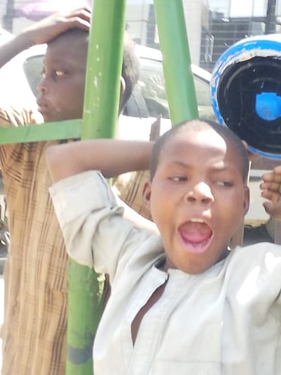 The image depicts two young males in an outdoor setting within the city of Gandu Albasa, Nigeria. In the foreground, a boy with short dark hair is captured mid-action, with his mouth wide open as if yawning or shouting. He is wearing a light-colored, possibly beige or off-white, collared shirt, unbuttoned at the top. His left arm is raised, with the hand positioned behind his head. Partially visible behind him and to his left is another boy, also with short dark hair, wearing a brown plaid or striped long-sleeved shirt. This second boy is looking towards the left side of the frame, with his right hand placed on top of his head. Two prominent vertical green poles run through the midground, separating and framing the subjects. To the right of the foreground boy's head, a blue and black object, appearing to be a plastic container or worn helmet, is visible. The background shows blurred outlines of structures, including a white vehicle and buildings, indicating an urban or semi-urban environment under daylight conditions.