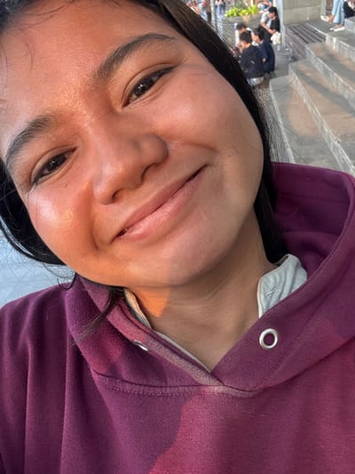 A close-up selfie shows a young woman with dark hair smiling at the camera. She is wearing a deep purple hooded sweatshirt with a light grey collar. The background is blurred, but appears to be an outdoor public space with tiered seating, possibly stairs or bleachers. In the distance, several people are visible, some sitting and others standing. The lighting suggests it is late afternoon or early evening, with the sun casting a warm glow on the woman's face. The setting is likely Ancol Beach City, North Jakarta, Indonesia, a popular recreational area with boardwalks and entertainment venues.