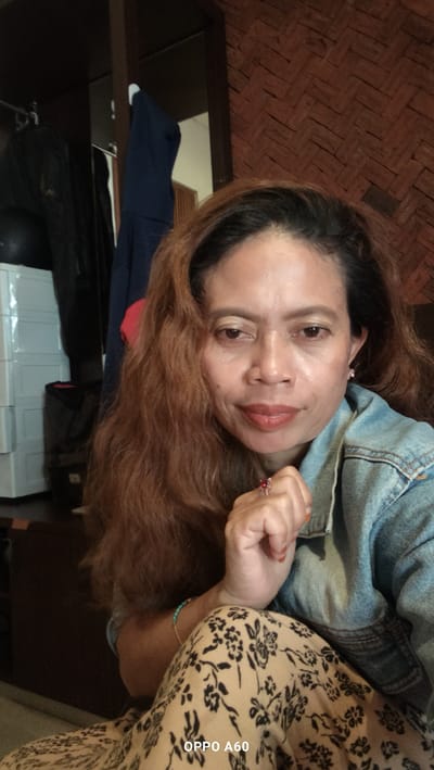 An Indonesian woman is captured in a close-up, selfie-style photograph, her gaze directed towards the camera. She has long, wavy brown hair and is wearing a light blue denim jacket over a patterned dress. Her left hand is raised, resting against her chin, adorned with a ring featuring a red stone. She wears a bracelet on her left wrist. The background suggests an indoor setting with wooden paneling on the right side and a wardrobe or closet on the left containing clothing. The location is identified as North Kuta, Indonesia, likely a residential area given the indoor environment and personal belongings. The lighting is natural and soft, illuminating the subject's face and creating subtle shadows. The overall impression is a casual, personal moment captured by the individual herself.