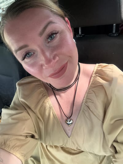The media file is a close-up, low-angle portrait of a fair-skinned woman with blonde hair, likely taken outdoors during the daytime, given the lighting. She is wearing a light tan, off-the-shoulder top with gathered elastic at the neckline and sleeves. A thin black cord necklace with a silver heart-shaped pendant is visible around her neck. Her makeup includes defined eyebrows, mascara, and a subtle lip gloss. Her blue eyes are looking slightly away from the camera. The background is blurred but appears to be a dark, textured surface, possibly car seats, and a bit of a dark metallic object. There is no visible text. The overall impression is a casual, possibly candid, shot, with the woman appearing relaxed and comfortable. The provided context of "Veliky Novgorod, Russia" is not discernible from the image itself.