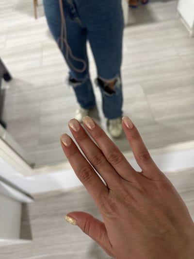 This media file shows a close-up view of a person's hand with glittery, light-colored nail polish. The hand is positioned in the foreground, with the fingers slightly curled. The background is out of focus, but appears to be an indoor setting with a light-colored, possibly tiled floor.
In the middle ground, a person's legs are visible, wearing ripped blue jeans. A thin, light-colored string or strap is draped across the jeans. The person is wearing sneakers.
The lighting suggests it is daytime. The overall scene appears casual, possibly taken in a home or a salon. There is no visible text in the image. The location context provided is Haskovo, Bulgaria.