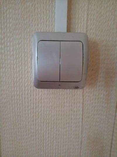 An indoor close-up shot shows a double light switch mounted on a textured, light-colored wall. The switch is silver-colored and has two rectangular buttons, one to the left and one to the right, with a slight gap between them. A portion of a white vertical piece of trim is visible above the switch. The wall has a repeating pattern of small, raised diamond shapes. The image does not contain any contextual information to determine its location within the city of Vilyuysk, Russia.