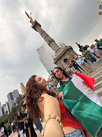 The image depicts a group of people gathered at the Monumento a la Independencia in Mexico City. In the foreground, two women are prominent. One woman, with long, curly brown hair, is holding a bracelet with yellow beads and has paint, possibly face paint, on her cheek. The other woman is holding a Mexican flag, which is predominantly green, white, and red, with the Mexican coat of arms visible. She is wearing a green and white jersey.
In the background, the Monumento a la Independencia, also known as El Ángel, stands tall with its golden sculpture of the Winged Victory of Samothrace at the top. The monument is made of stone and features intricate carvings and statues at its base. Several other people are visible, some wearing green jerseys, suggesting a gathering related to a sporting event or national celebration. Behind the monument, modern buildings are visible, including a skyscraper with a logo that appears to be for HSBC. The sky is overcast with clouds. The scene appears to be an outdoor public gathering in a city square.
