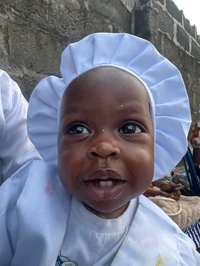 The image captures a young child with dark skin, positioned in the foreground and looking directly at the camera with wide-open, dark eyes. The child wears a white, pleated or ruffled bonnet and a white long-sleeved outer garment, which appears to be layered over another white top. Small, reflective, glitter-like specks are visible on the child's forehead and left cheek. A light reddish-pink stain is present on the upper left shoulder area of the outer white garment, while a yellowish stain is seen on the lower right front of the same garment. The child's mouth is slightly open, revealing two lower and two upper front teeth.
The background consists of a rough, grey wall constructed from what appears to be concrete blocks or large stones, showing distinct textures and mortar lines, particularly prominent behind the child's head and to the upper right. The top edge of this wall is jagged. To the immediate left of the child, another person's white garment is partially visible, suggesting close proximity. To the right of the child, blurred elements indicate the presence of other individuals and objects: a darker-skinned hand holding a black smartphone, a beige, fuzzy object, and blue and white striped fabric. The scene is situated in Badagry, Nigeria.