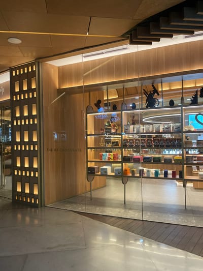 The image depicts a store with a modern and clean aesthetic. The primary subjects are shelves filled with various chocolate boxes and decorative items, suggesting a retail environment focused on confectionery. The scene is set indoors, with warm lighting emanating from behind wooden panels and illuminated squares on a lower wall, creating a cozy and inviting atmosphere. The display shelves are made of glass and metal, showcasing an array of colorful chocolate packaging, some of which are arranged in neat rows.
Notable details include what appears to be a sign visible through the glass with the text "THE RR CHOCOLATE" printed vertically. The reflection in the glass also hints at the presence of a display with a light blue glow and some decorative elements. The overall impression is that of a well-lit, upscale chocolate shop. The time of day is not explicitly indicated but the interior lighting suggests it could be evening or a dimly lit indoor space. The location cue "Kuta, Indonesia" places this within a specific commercial area. There are no people visible in the image, and the scene is static, focusing on the products and the store's interior design.