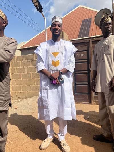 Two men are standing outdoors in Rigasa, Nigeria. One man is wearing a white, patterned robe with gold embroidery, a white cap, and beige shoes. He is holding a smartphone and a purple object. The other man is wearing a tan outfit with a matching hat and black shoes. Behind them are a building with a brown door and a red roof, a brick wall, and a dirt ground. A streetlight is visible in the background.