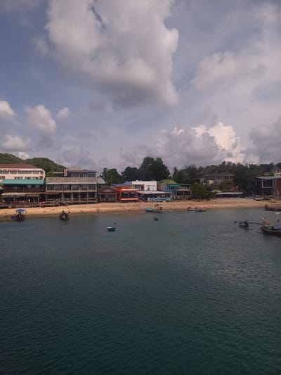 The image depicts a coastal scene in Tambon Ko Tao, Thailand. In the foreground, dark blue water with gentle ripples fills the lower portion of the frame, with several boats anchored or drifting. A sandy beach stretches along the midground, lined with various buildings and structures, including a hospital, restaurants, and accommodations. Some buildings have a veranda and are located near the beach. A few people are visible on the beach and near the water. The background is filled with lush green vegetation and trees, adding to the tropical setting. Above, a partially cloudy sky dominates the upper part of the image.