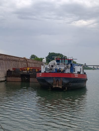 A cloudy, overcast day shows a large, red and white vessel docked in what appears to be a harbor or river in Šabac, Serbia. The vessel, labeled "SPIRIT," has a black hull and a prominent red bow with two large anchors. The vessel's superstructure is white with blue accents and features various equipment, including radar domes and ventilation units. To the left of the vessel is a large, concrete structure, possibly a seawall or dock. A dark-colored barge with Cyrillic lettering that reads "ЛАКО 12" is moored next to the concrete structure. The water is rippled, reflecting the overcast sky. In the background, trees and a bridge can be seen on the horizon, suggesting an urban or semi-urban environment. No people are visible in the image.